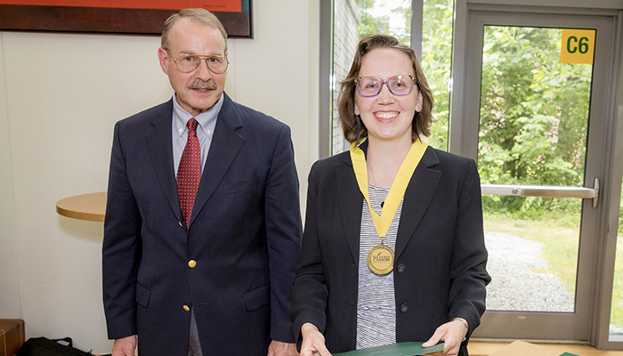 A man in a blue blazer stands next to a woman in a dark jacket and striped skirt wearing a gold medal.