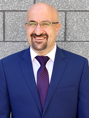 A bald man with a short beard in a blue jacket and purple tie smiles.