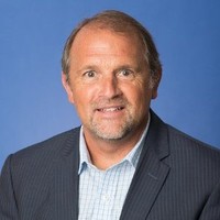 A man in a dark jacket and checked shirt with a stubble beard looks at the camera.