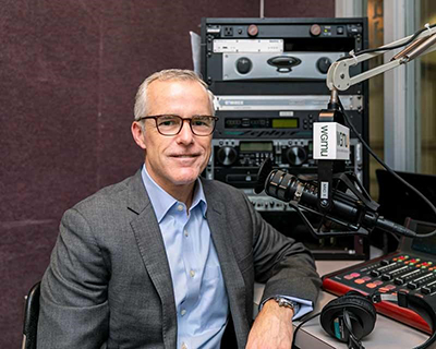 A man in eyeglasses and a gray suit jacket and blue shirt sits in front of a microphone.
