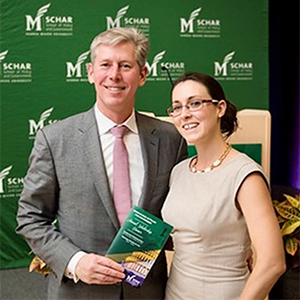 A man in a gray suit stands next to a woman in a beige dress as they smile at the camera.