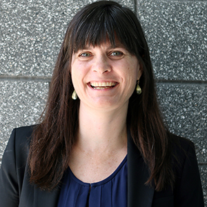 A woman with dark hair and bangs in a black jacket smiles broadly at the camera.