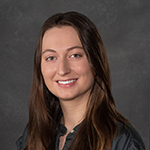 A woman in a black shirt stands smiling in front of a gray background