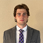A young man in a gray suit, white collared shirt, and tie stands and smiles for the camera