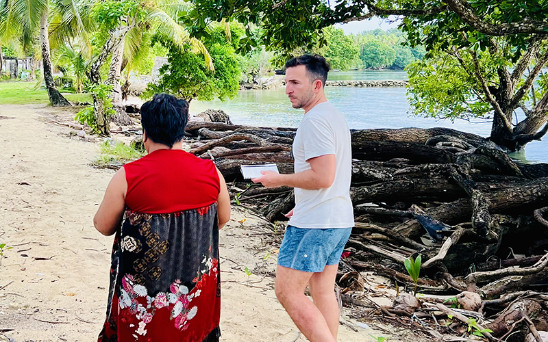 A man wearing a T-shirt, shorts, and flip-flops walks along a beach with a woman wearing a red and black dress. 
