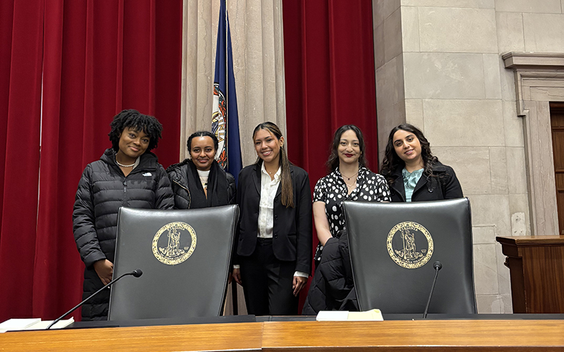 Five young women stand behind two large chairs and a long wooden desk in a courtroom.