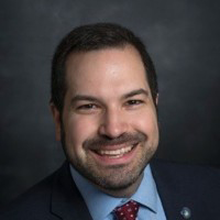 A man with a beard wears a black suit, blue collared shirt, and red tie, stands and smiles