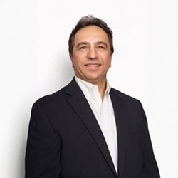 A man in a black suit and white collared shirt stands and smiles
