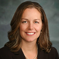 A woman in a black shirt stands in front of a gray background and smiles