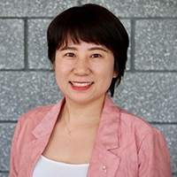 A woman in a pink jacket and white shirt stands in front of a cinder block wall and smiles