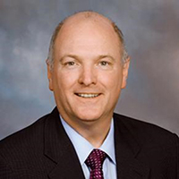 A man in a black suit, blue shirt, and red tie, stands and smiles