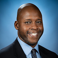 A man in a black suit, blue shirt, and tie stands in front of a blue background and smiles