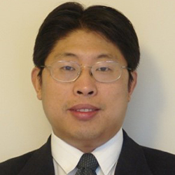 A man wearing glasses, a black suit, white collared shirt and tie stands and smiles